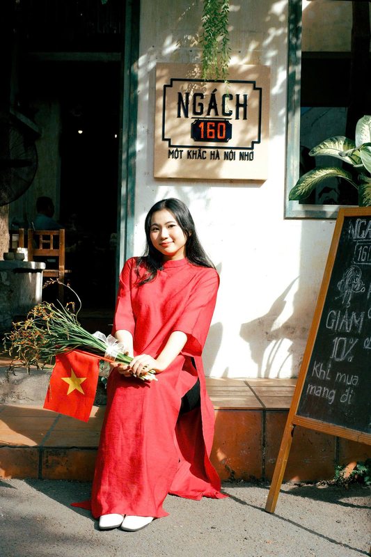 A young woman in a red ao dai holding flowers and a small Vietnamese flag, seated on the step of a cafe beside its chalkboard menu