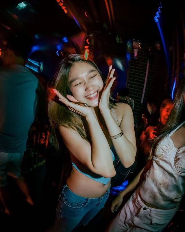 A young woman laughing with her hands to her cheeks on a crowded dance floor lit in pink and blue