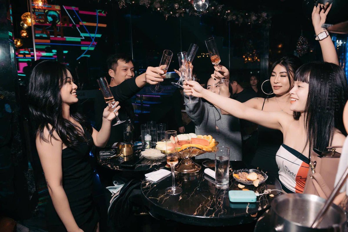 A group of friends raising champagne flutes in a toast over a fruit platter at a lounge table under neon lights