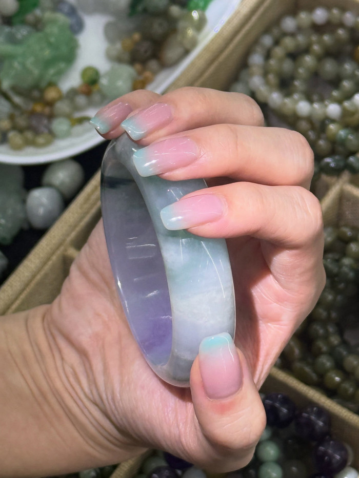 A lavender-green jade bangle held up in the salon, trays of stones behind