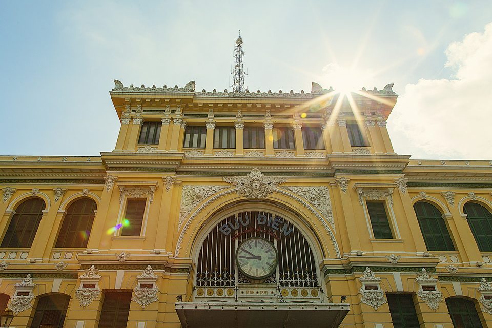 The Central Post Office photographed from below, sunlight flaring past the clock and the words BƯU ĐIỆN carved above the entrance.