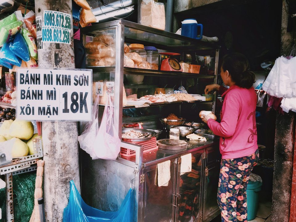 A street bánh mì cart with its price board reading 18K, the vendor filling a baguette to order beside stacked bread and fillings.