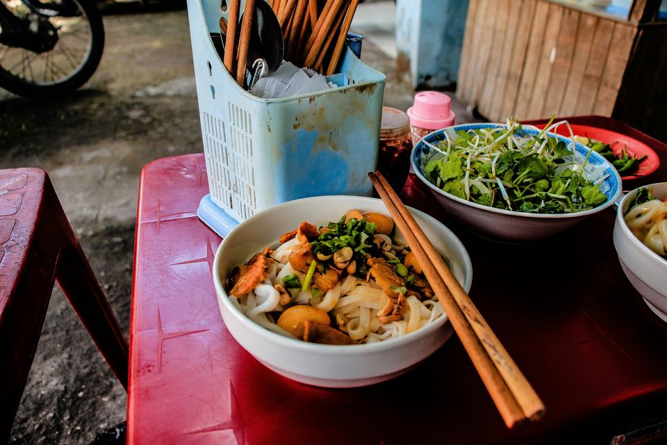 A bowl of rice noodles on a red plastic table at a pavement stall, with a shared plate of herbs and bean sprouts and a caddy of chopsticks beside it.