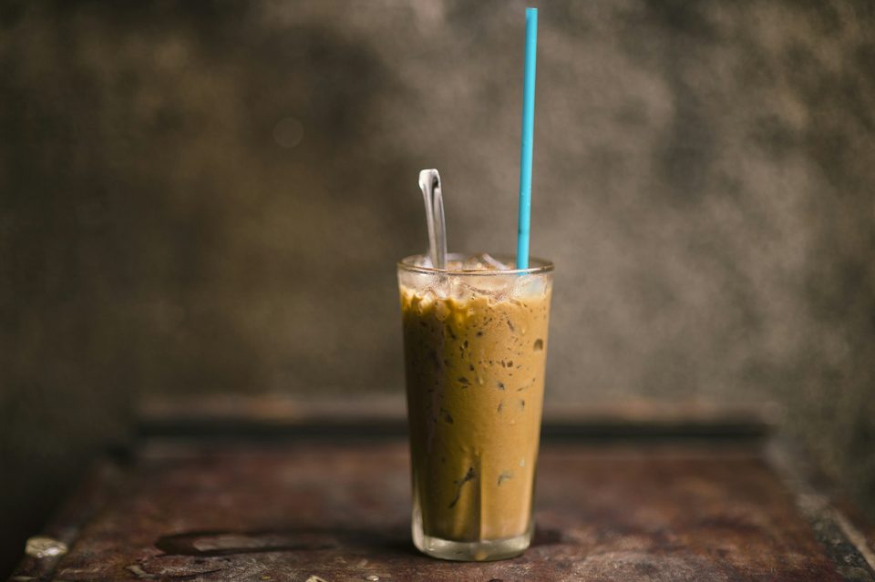 A tall glass of iced Vietnamese coffee, the condensed milk still settling through it, a long spoon and straw standing in the glass.