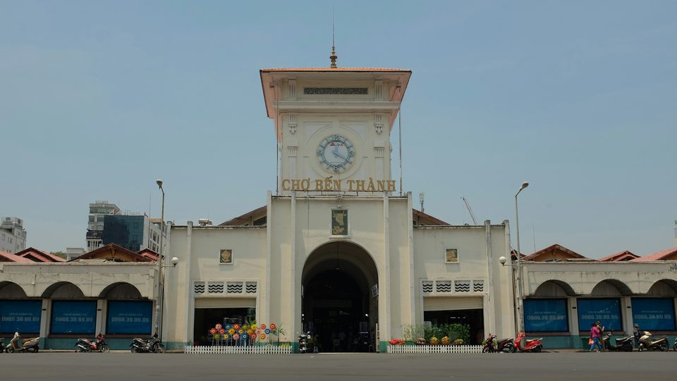 The clock tower entrance of Bến Thành Market, CHỢ BẾN THÀNH in gold lettering beneath the clock, motorbikes parked along the front.