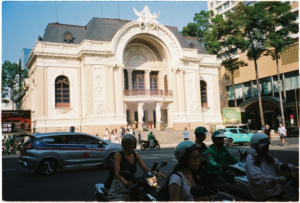 The Saigon Opera House in full sun, its ornate facade above a street of motorbike riders and a taxi waiting at the kerb.