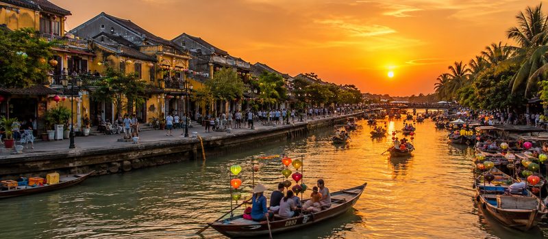 A river at sunset with wooden boats carrying lit paper lanterns, low tiled shophouses along one bank and palms along the other.