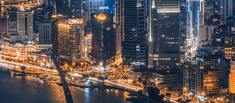 A dense city skyline at night, lit towers above a bright waterfront strip and a cable-stayed bridge crossing the river below.